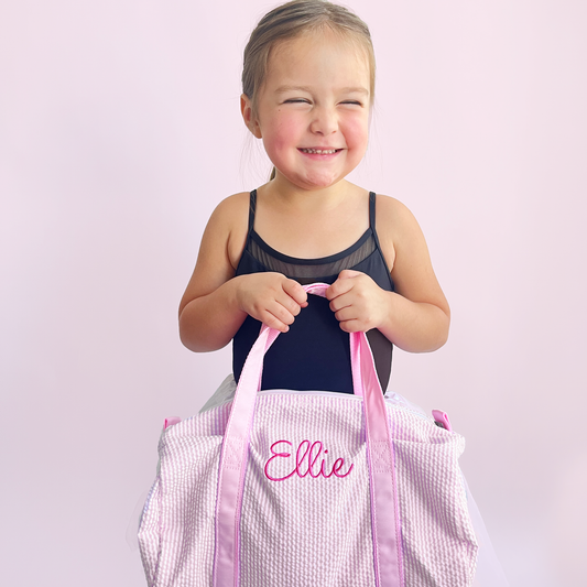 Little girl wearing a ballet outfit and holding a mini pink seersucker duffle bag with a custom name embroidered in a cute script font and pink thread.