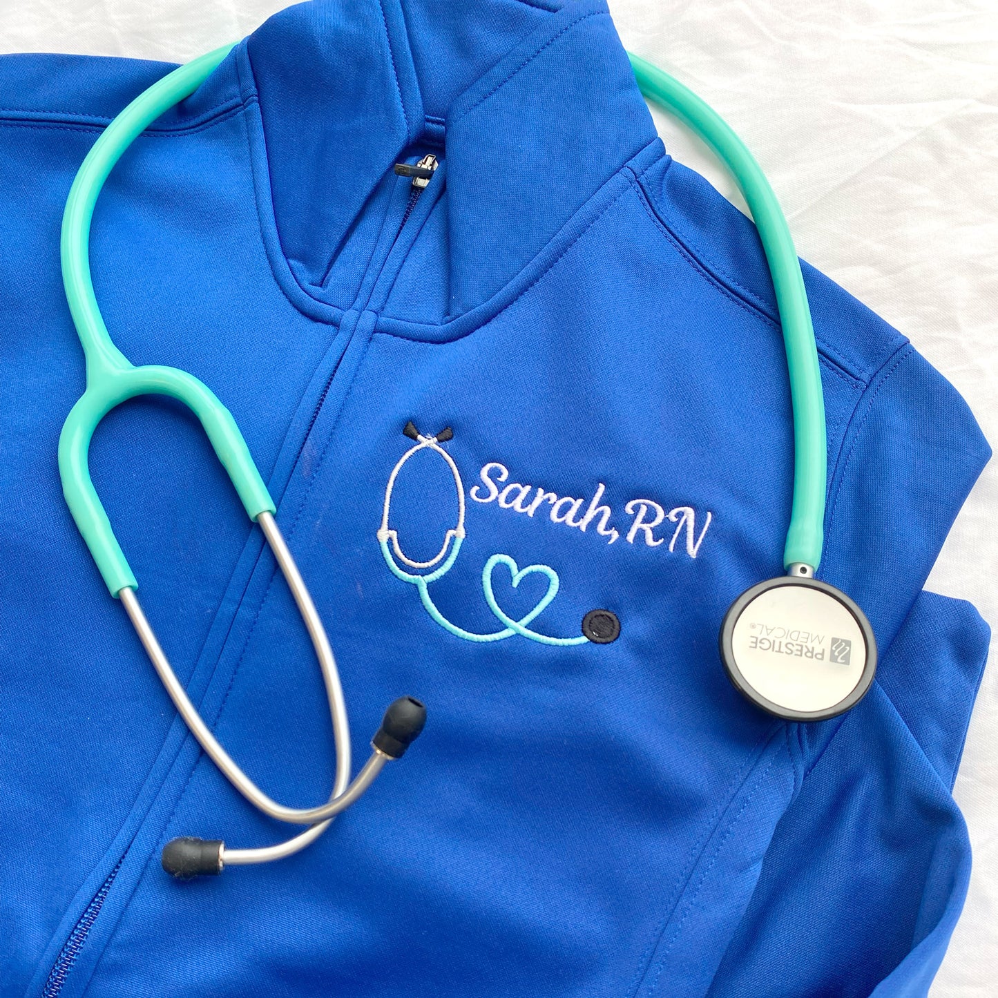 close up of a royal blue full zip polyester nurse jacket with custom embroidered heart stethoscope and name in white and sky thread!