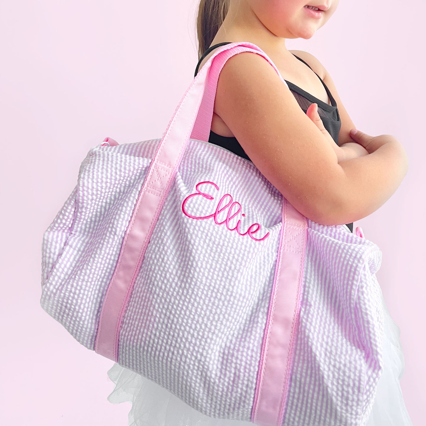 Little girl wearing a ballet outfit and holding a mini pink seersucker duffle bag with a custom name embroidered in a cute script font and pink thread.