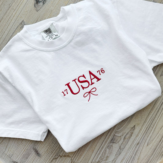 white comfort colors t-shirt with custom 1776 USA dainty bow embroidered design on the center chest in red thread