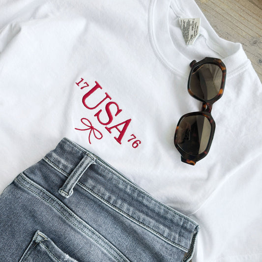 white comfort colors t-shirt with custom 1776 USA dainty bow embroidered design on the center chest in red thread paired with sunglasses and jeans