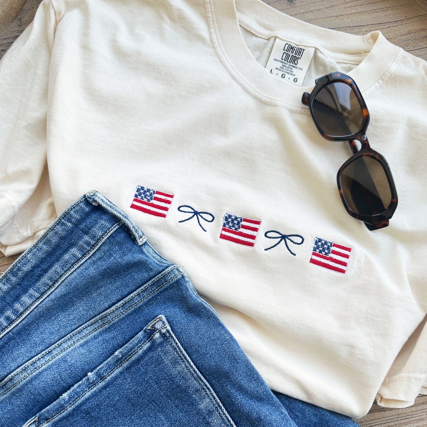 ivory comfort colors tee with an alternating row of mini american flags and dainty bows embroidered on the center chest, paired with sunglasses and jeans