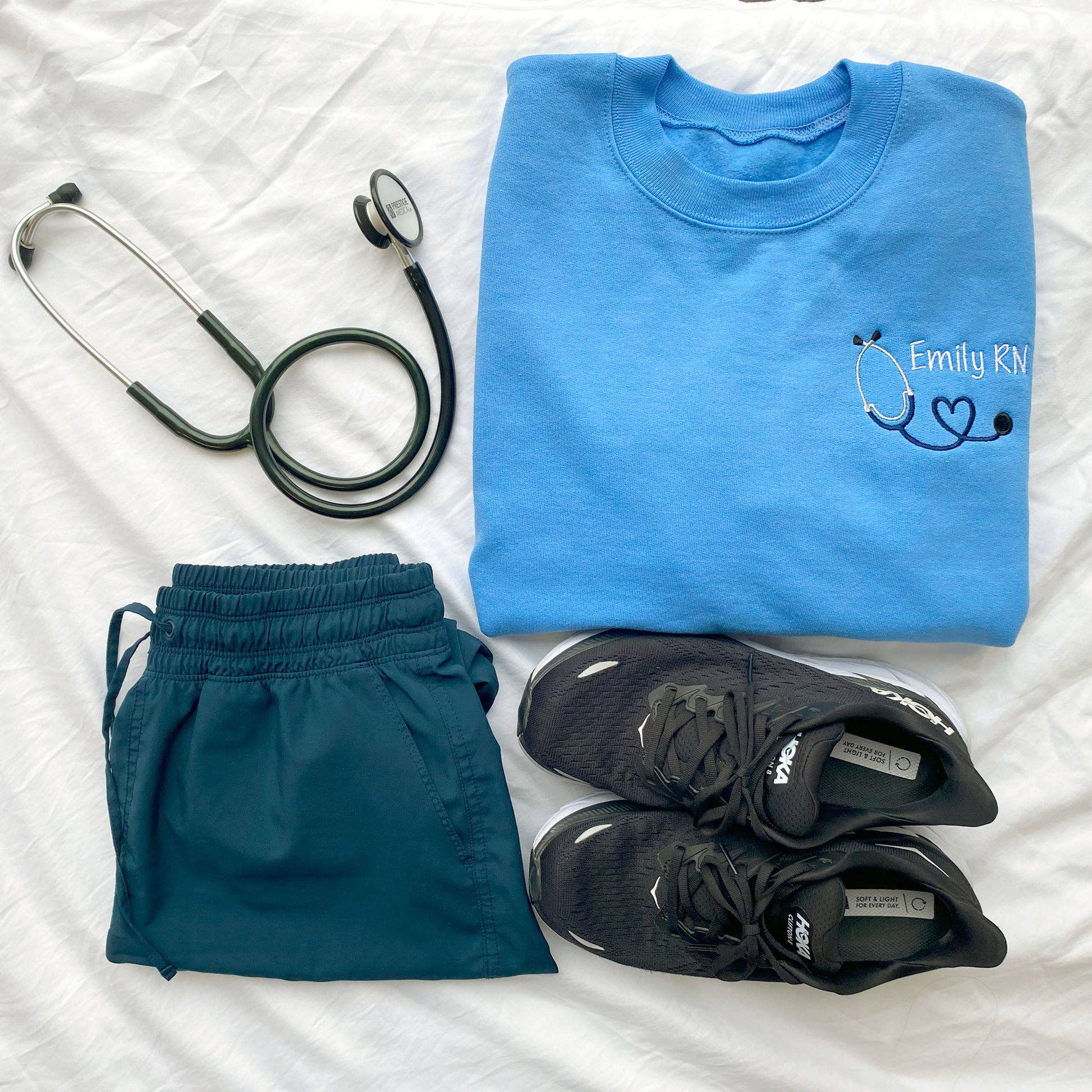 styled flat lay of a  Carolina blue crewneck sweatshirt with custom mini heart stethoscope design embroidered on the left chest in navy and white threads with navy scrubs, sneakers, and a stethoscope.