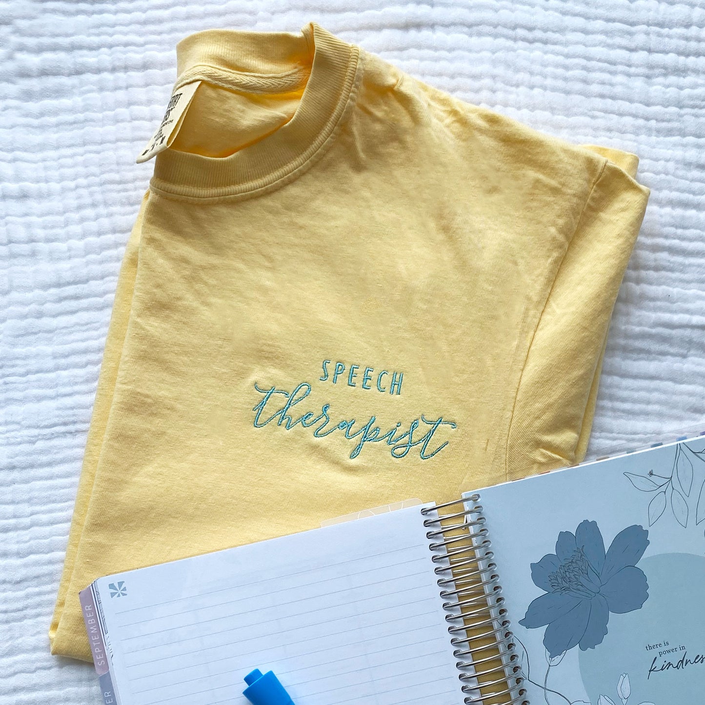 flat lay of a custom embroidered speech therapist design in sky blue thread on a butter yellow comfort colors t-shirt