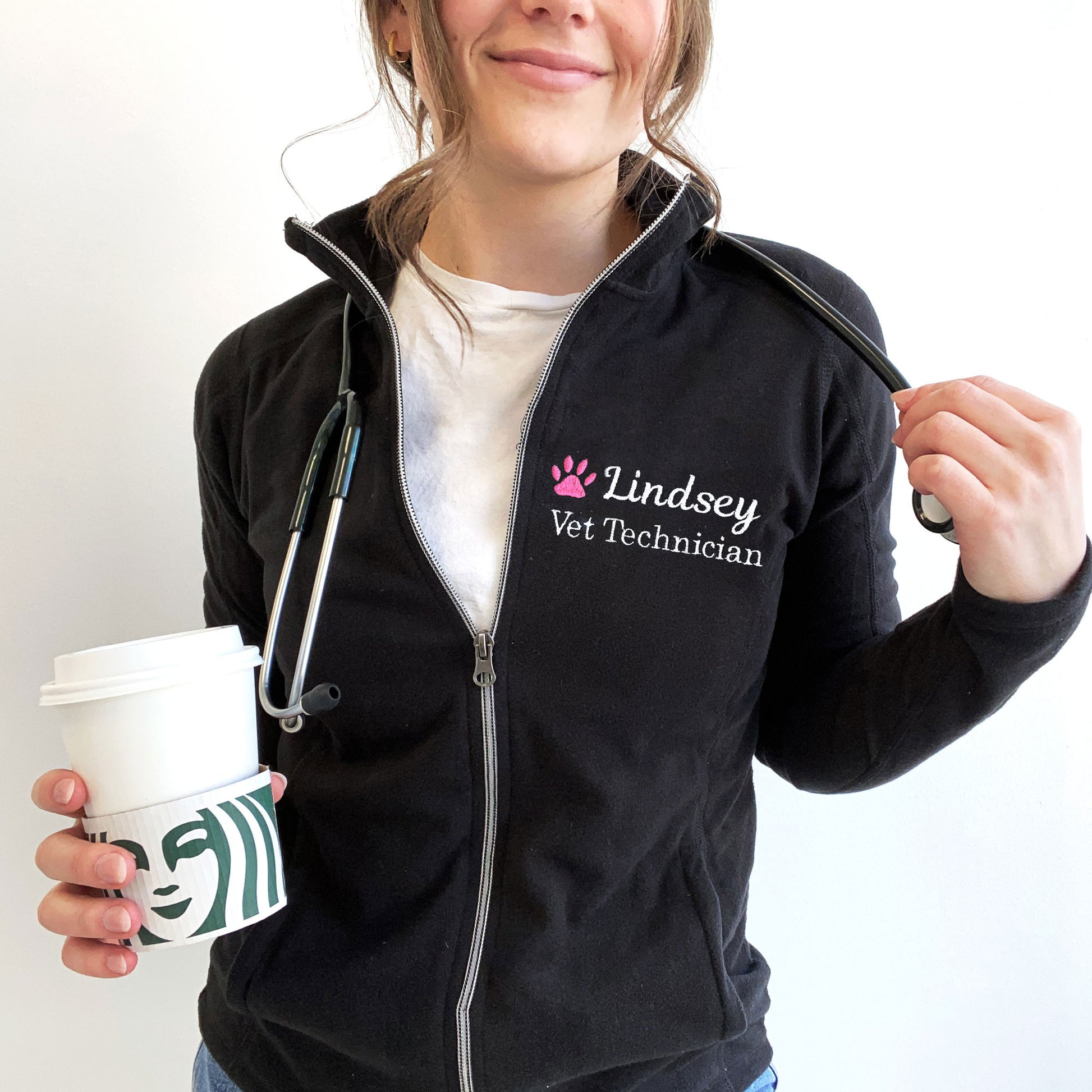 young woman wearing a black full zip fleece jacket with embroidered pink paw print and name Lindsey with Vet Technician embroidered on the left chest in white thread