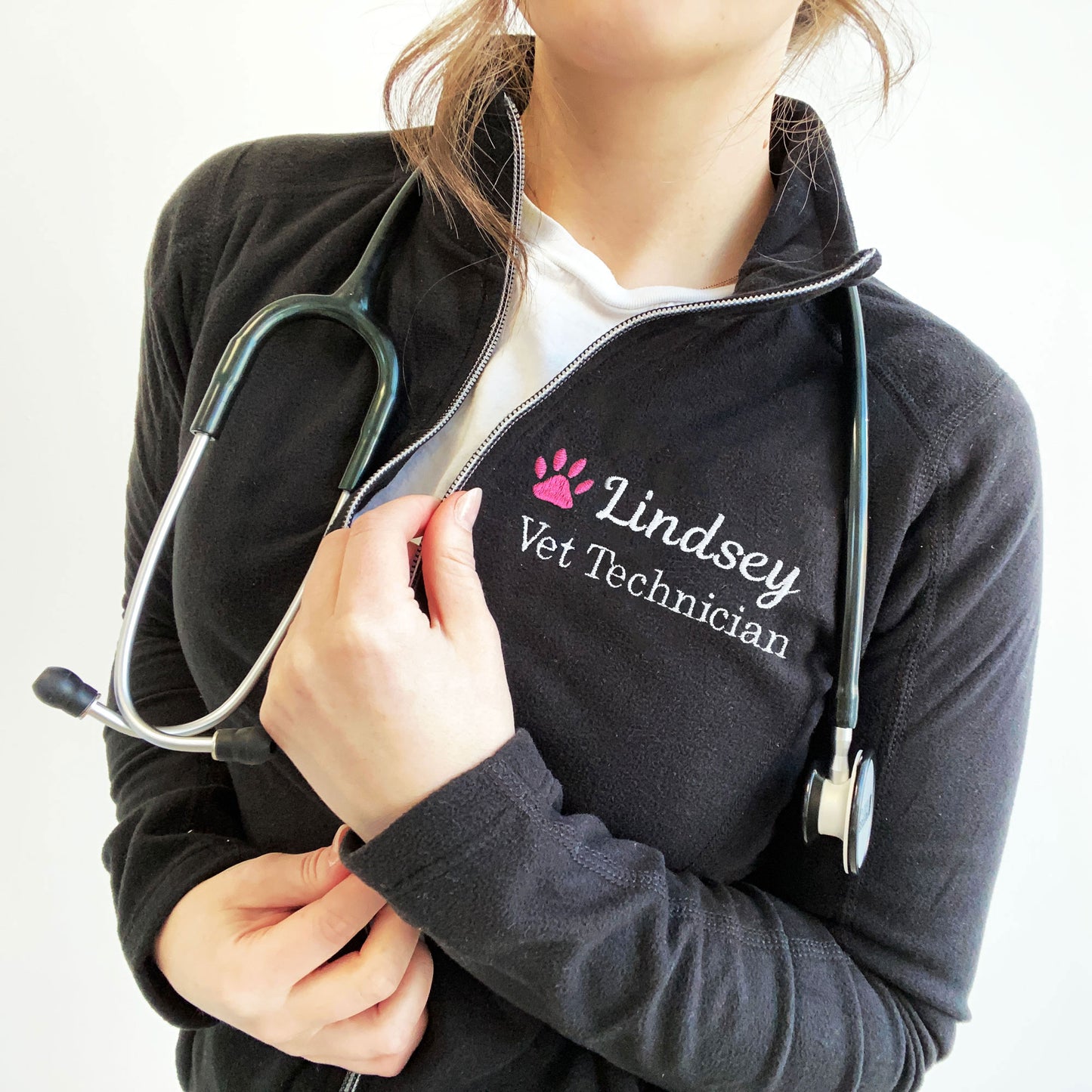 young woman wearing a black full zip fleece jacket with embroidered pink paw print and name Lindsey with Vet Technician embroidered on the left chest in white thread