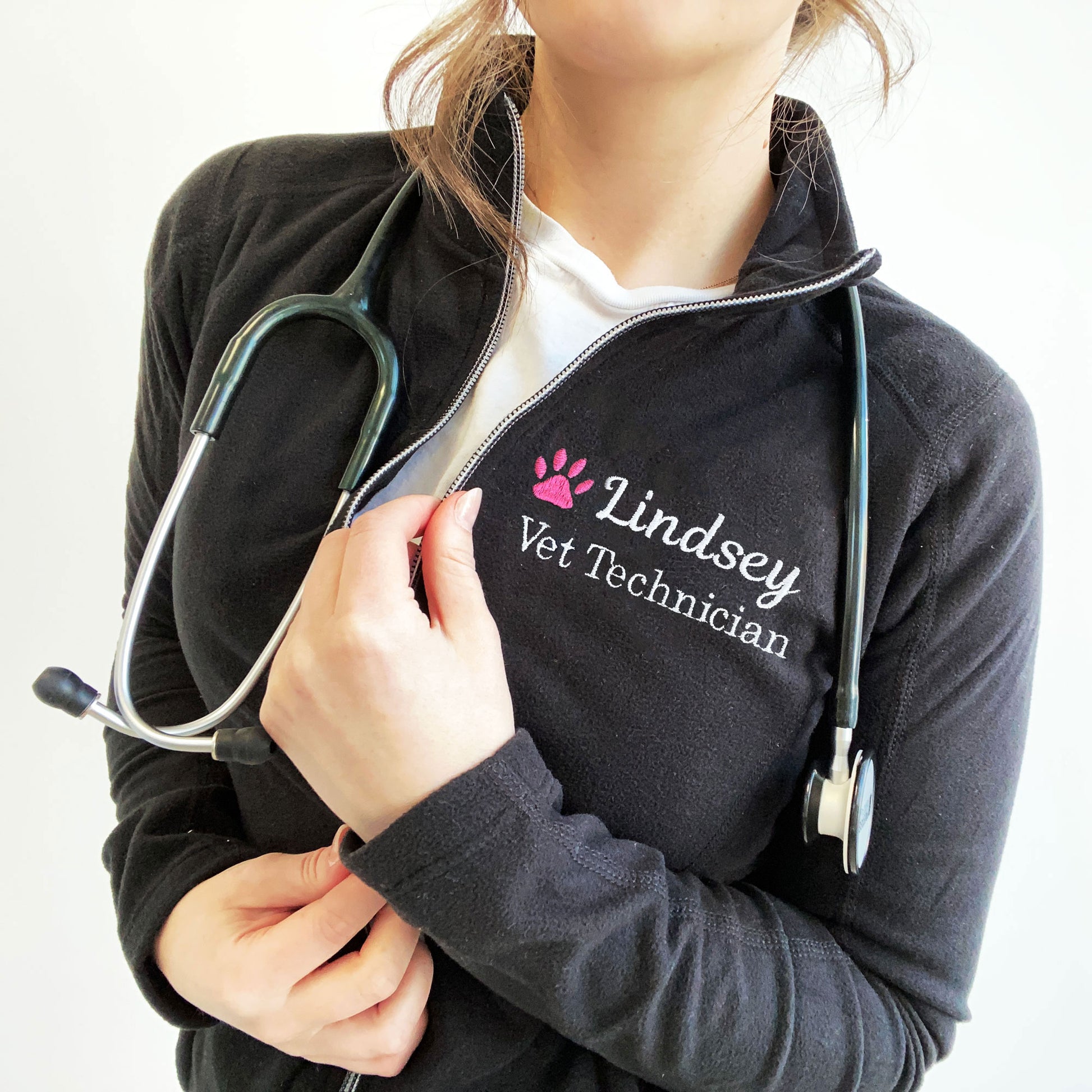 young woman wearing a black full zip fleece jacket with embroidered pink paw print and name Lindsey with Vet Technician embroidered on the left chest in white thread