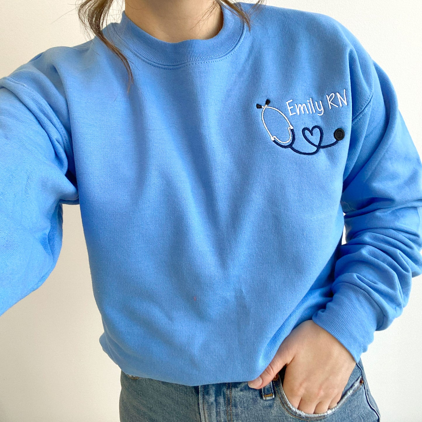young woman wearing a Carolina blue crewneck sweatshirt with custom mini heart stethoscope design embroidered on the left chest in navy and white threads