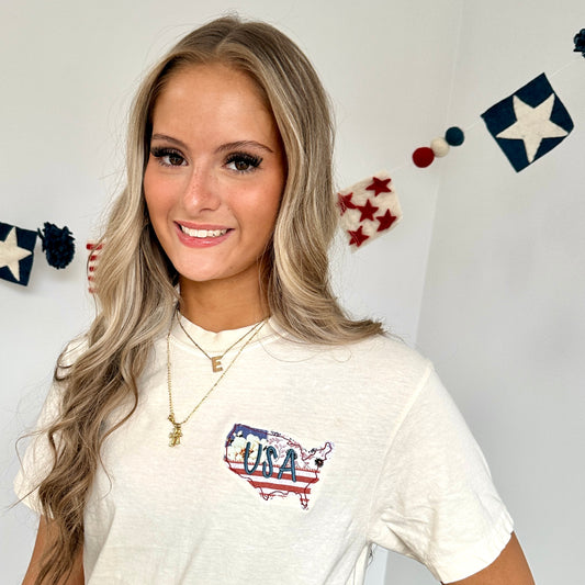 girl standing in front of a white wall with a patriotic banner hanging behind her. She's wearing an ivory comfort colors t-shirt with a custom usa outlined applique embroidery on the left chest. fabric is a multi-colored pattern with USA embroidered in blue thread over top