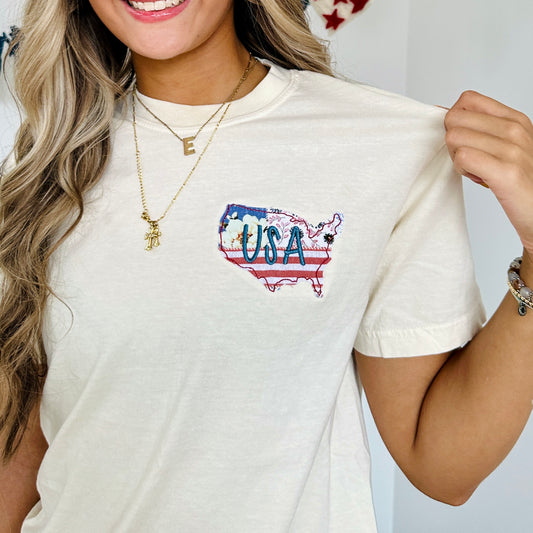 close up of a girl wearing an ivory comfort colors t-shirt with a custom usa outlined applique embroidery on the left chest. fabric is a multi-colored pattern with USA embroidered in blue thread over top