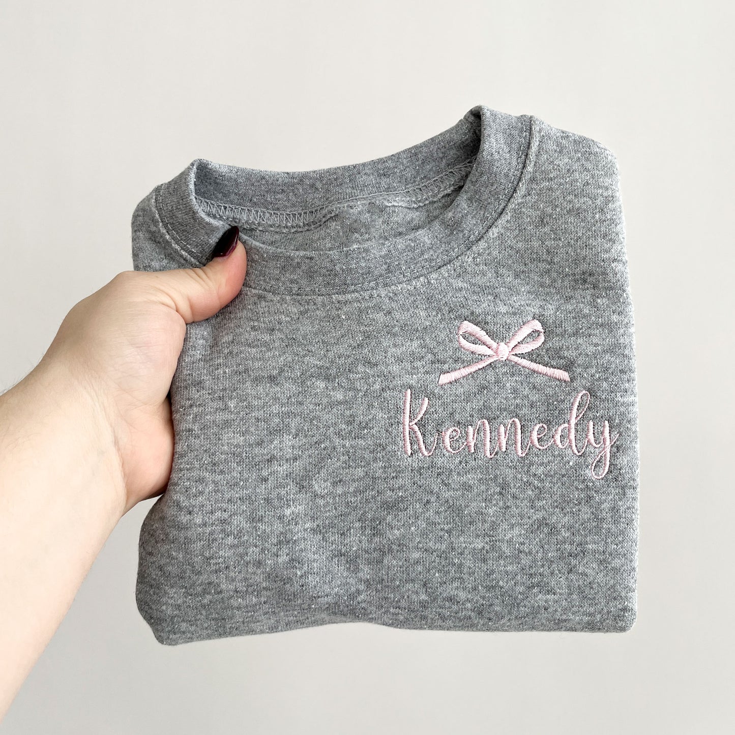 heather grey youth crewneck sweatshirt with embroidered bow and name design on the left chest in baby pink thread