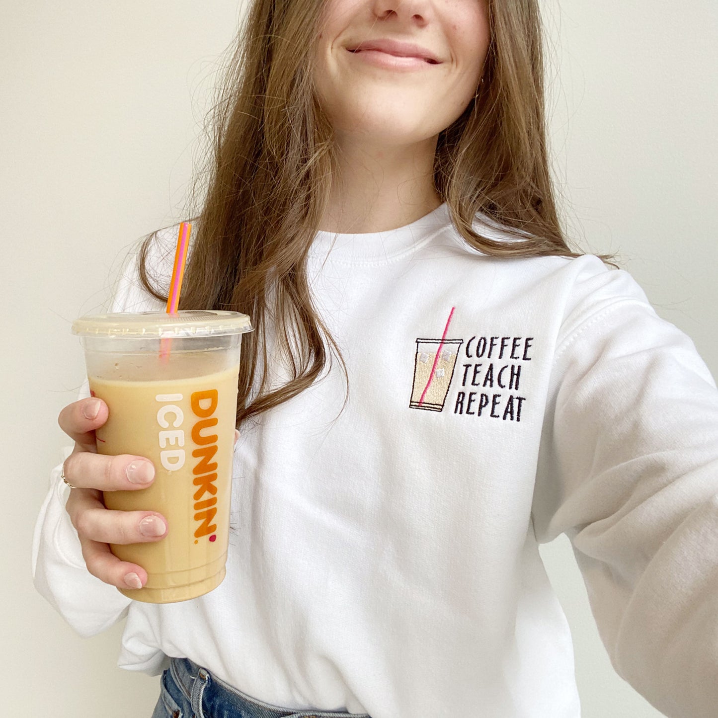 teacher wearing a white crewneck sweatshirt with a custom embroidered design on the left chest featuring an iced coffee design