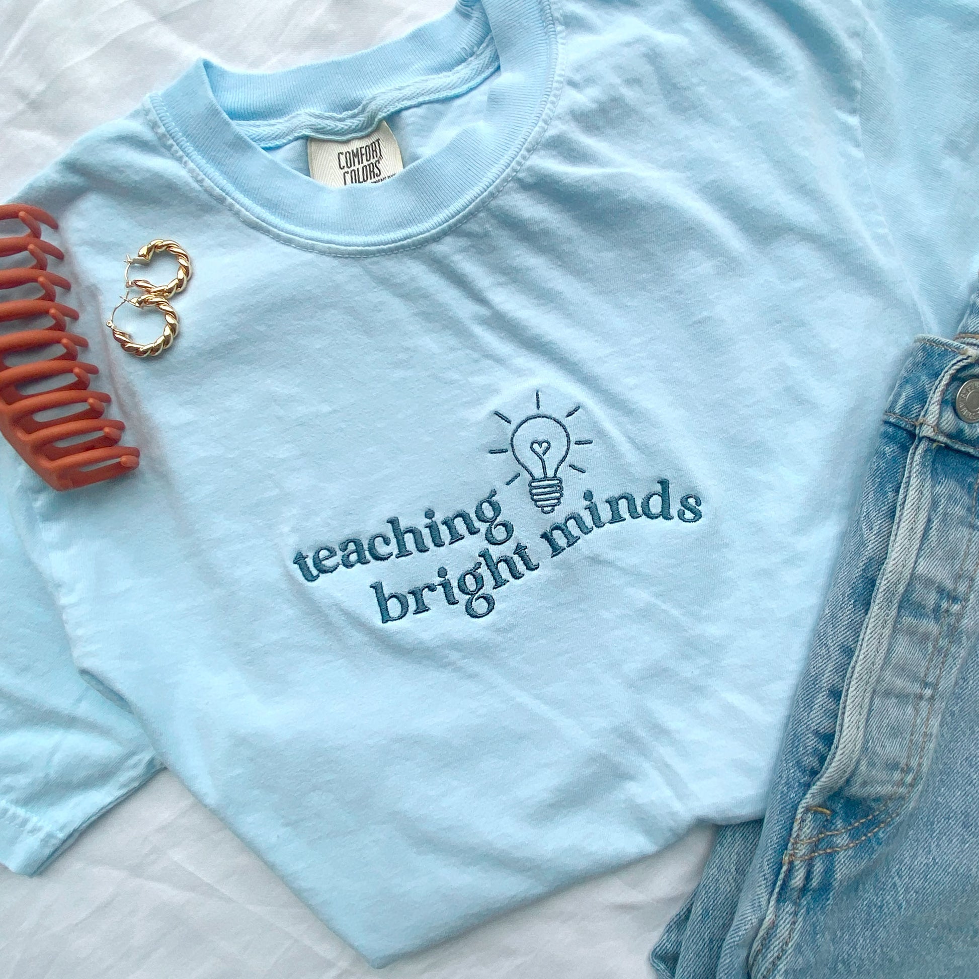 flat lay of a chambray comfort colors tshirt with teaching bright minds wavy text and cute heart lightbulb design embroidered across the chest in french blue threads styled with jeans, earrings, and a claw clip