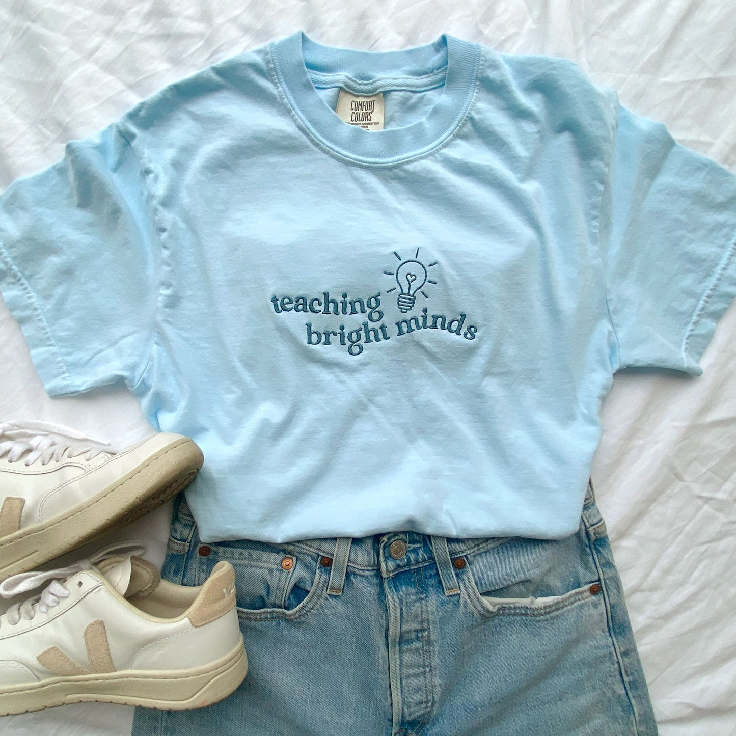 flat lay of a chambray comfort colors tshirt with teaching bright minds wavy text and cute heart lightbulb design embroidered across the chest in french blue threads styled with jeans and sneakers