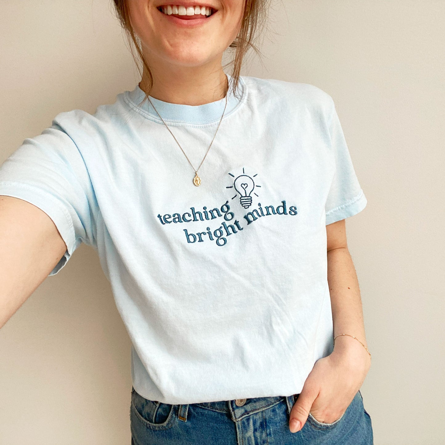 young woman modeling a chambray comfort colors tshirt with teaching bright minds and cute heart lightbulb design embroidered across the chest in french blue threads