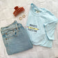 flat lay of a a chambray comfort colors tshirt with embroidered progress over perfection and pencil design styled with jeans, earrings, and a claw clip