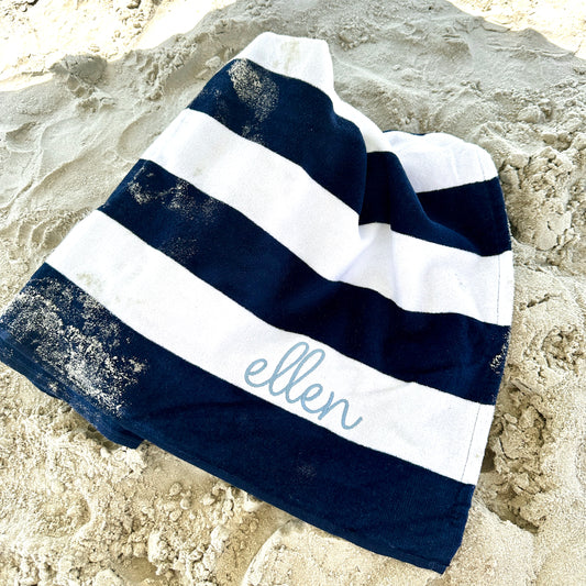 navy blue and white striped cabana beach towel with custom name embroidery in a cursive blithe font along the second stripe, horizontally. Embroidery is in powder blue thread. Towel is laying partially folded on a sandy beach