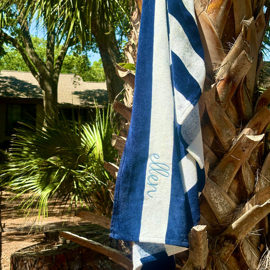 navy blue and white striped cabana beach towel with custom name embroidery in a cursive blithe font along the second stripe, horizontally. Embroidery is in powder blue thread. Towel is hanging off a palm tree