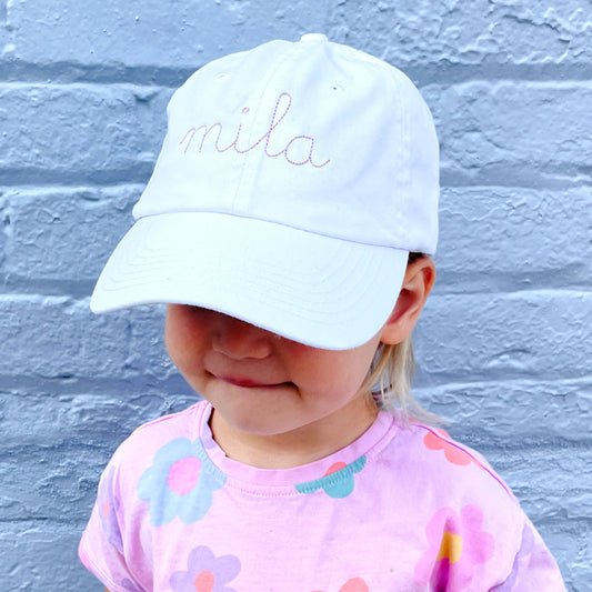 little girl wearing a youth white baseball hat with embroidered stitched script name in mauve thread