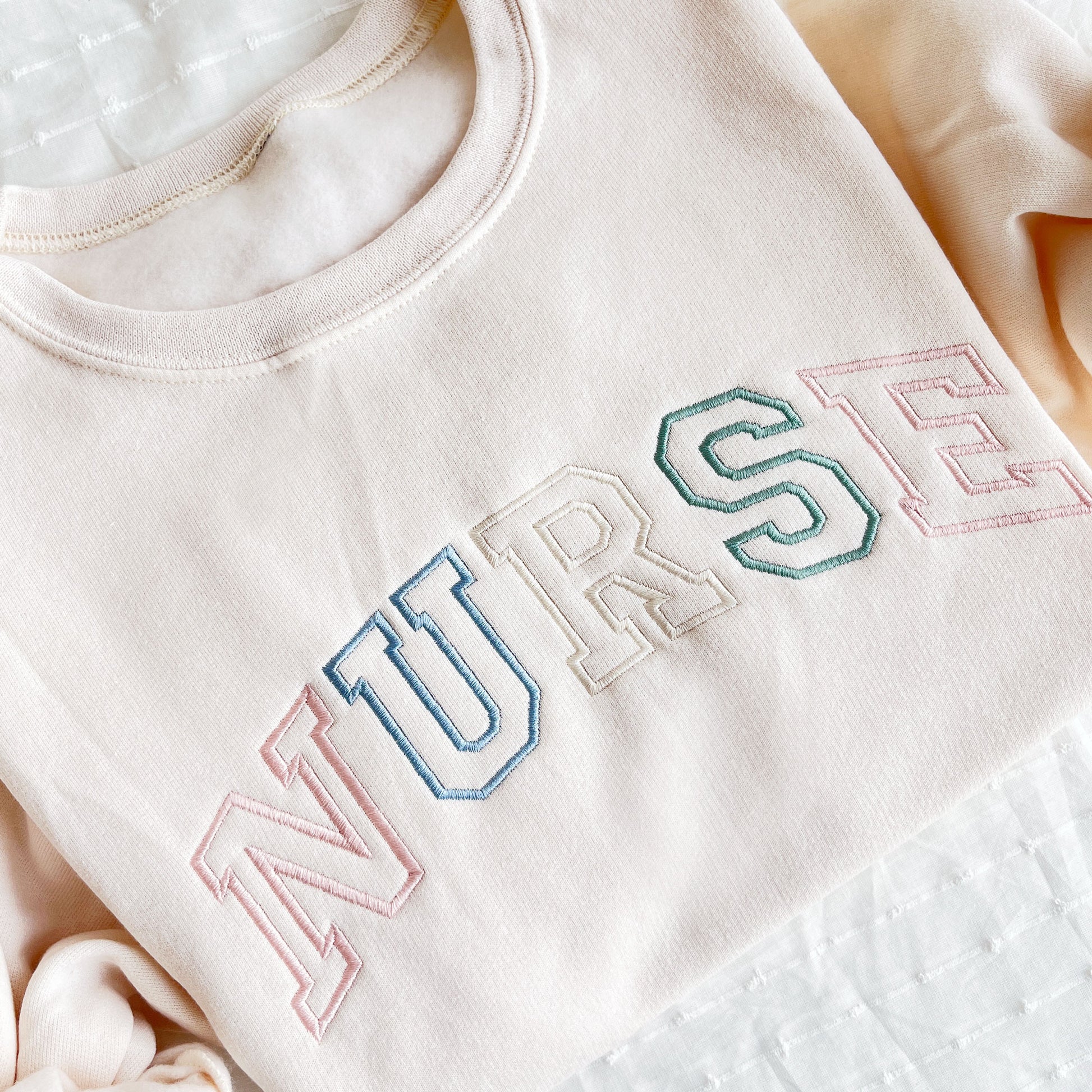 sweet cream crewneck sweatshirt with custom NURSE athletic outlined font embroidered in pastel pink, purple, natural, and eucalyptus thread