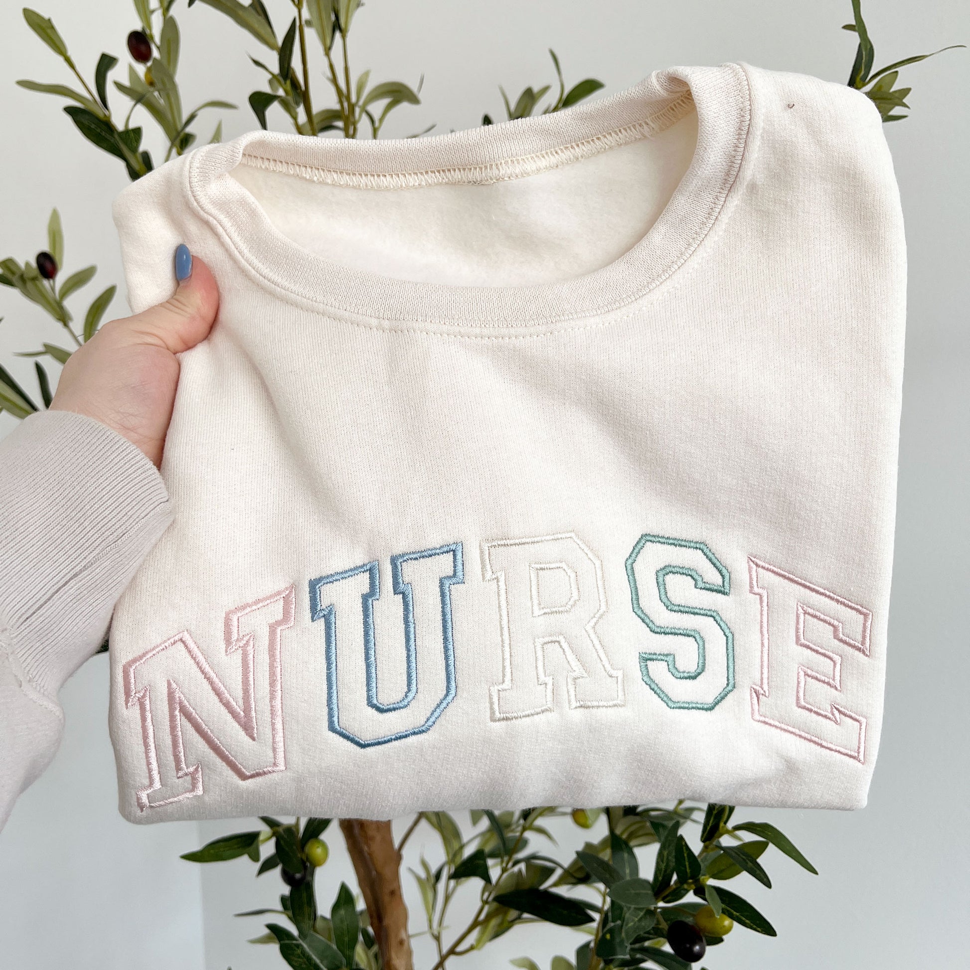 sweet cream crewneck sweatshirt with custom NURSE athletic outlined font embroidered in pastel pink, purple, natural, and eucalyptus thread