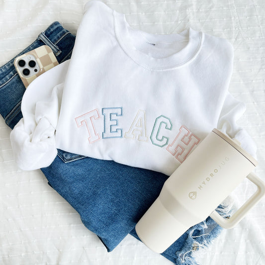 white crewneck sweatshirt with custom embroidered varsity outlined font embroidery reading TEACH in alternating pastel thread colors including: powder pink , powder blue, natural, and eucalyptus.