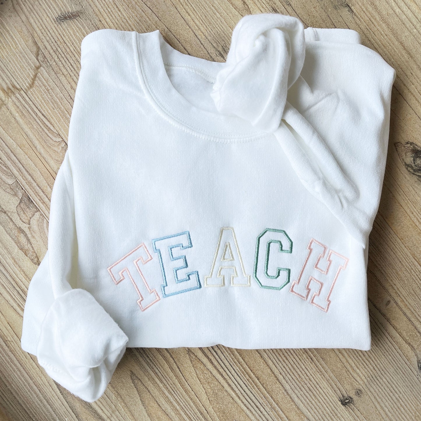 white crewneck sweatshirt with custom embroidered varsity outlined font embroidery reading TEACH in alternating pastel thread colors including: powder pink , powder blue, natural, and eucalyptus.