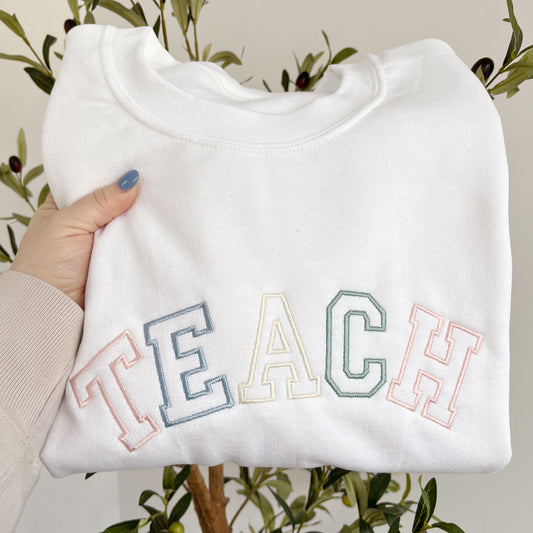 white crewneck sweatshirt with custom embroidered varsity outlined font embroidery reading TEACH in alternating pastel thread colors including: powder pink , powder blue, natural, and eucalyptus.