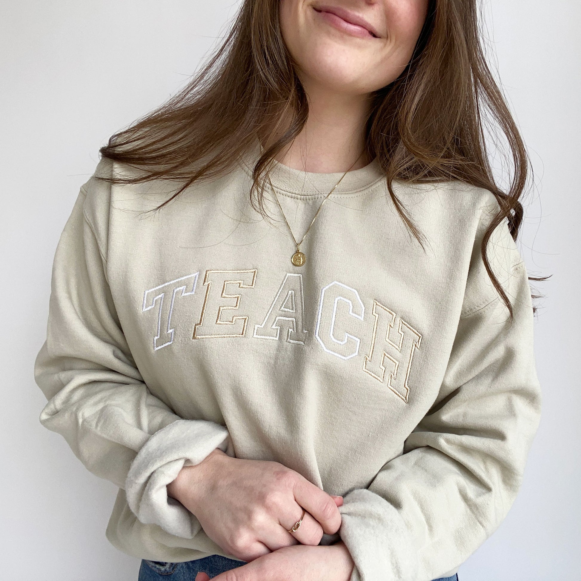 close up of a young woman wearing a sand crewneck sweatshirt with embroidered neutral color block teach design across the chest