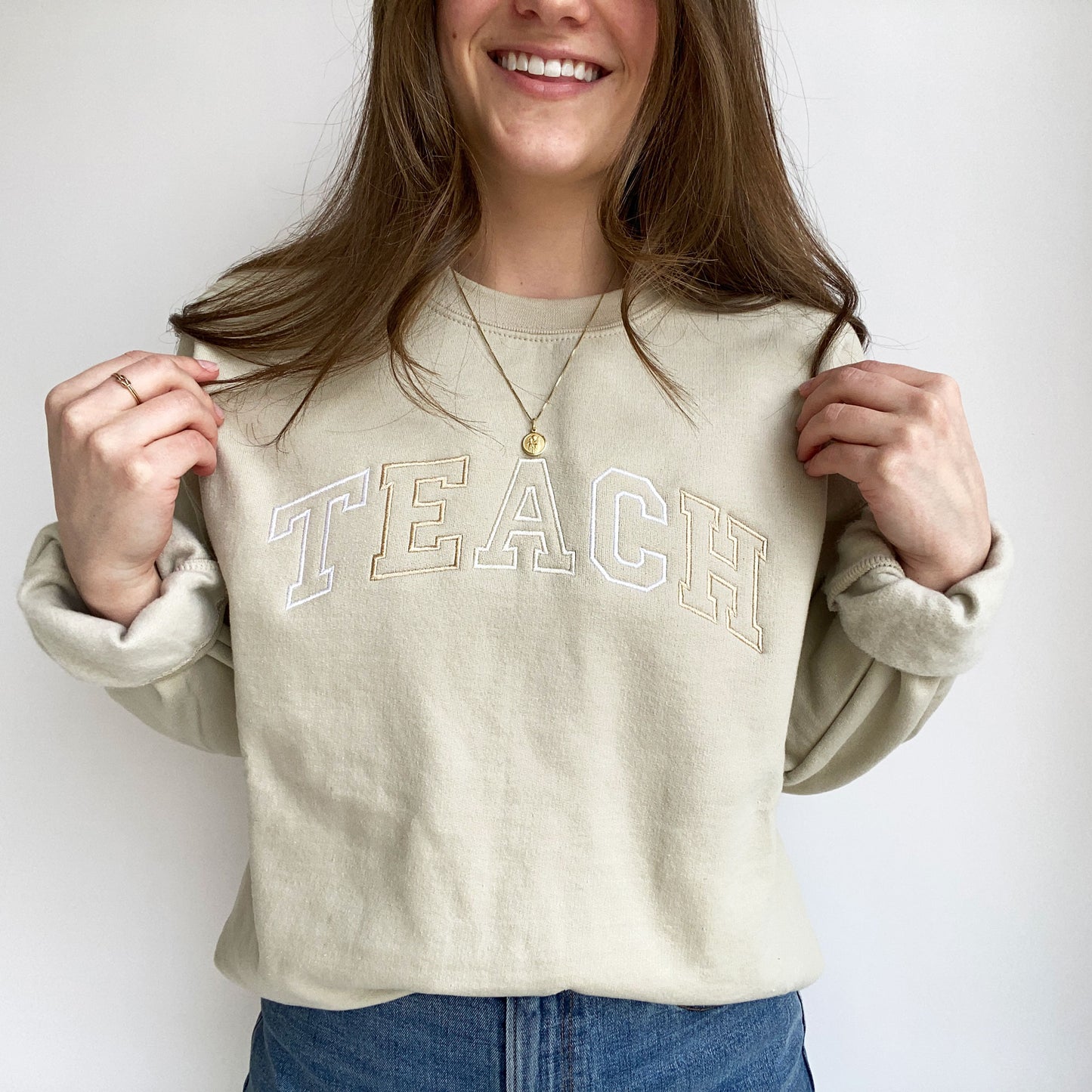 young woman wearing a sand crewneck sweatshirt with embroidered neutral color block teach design across the chest