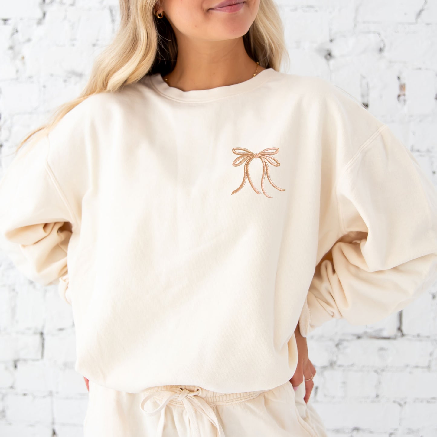 woman wearing an ivory comfort colors lightweight crewneck sweatshirt with embroidered ribbon bow on the left chest in camel thread