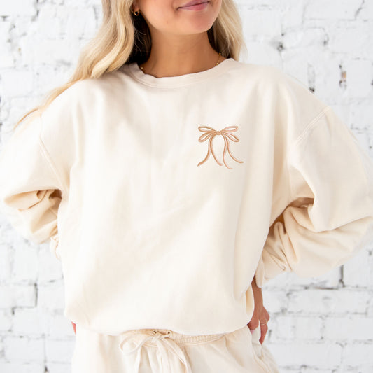 woman wearing an ivory comfort colors lightweight crewneck sweatshirt with embroidered ribbon bow on the left chest in camel thread