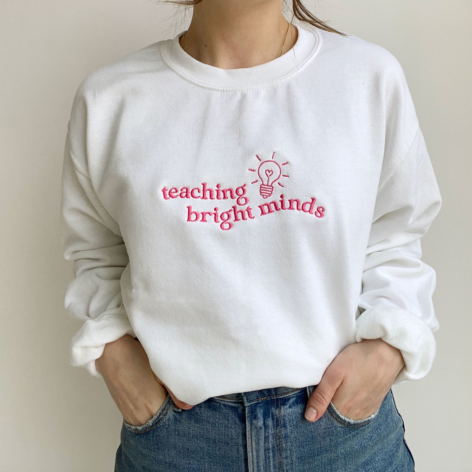 young woman wearing a white teacher pullover sweatshirt with a teaching bright minds lightbulb embroidered design on the chest