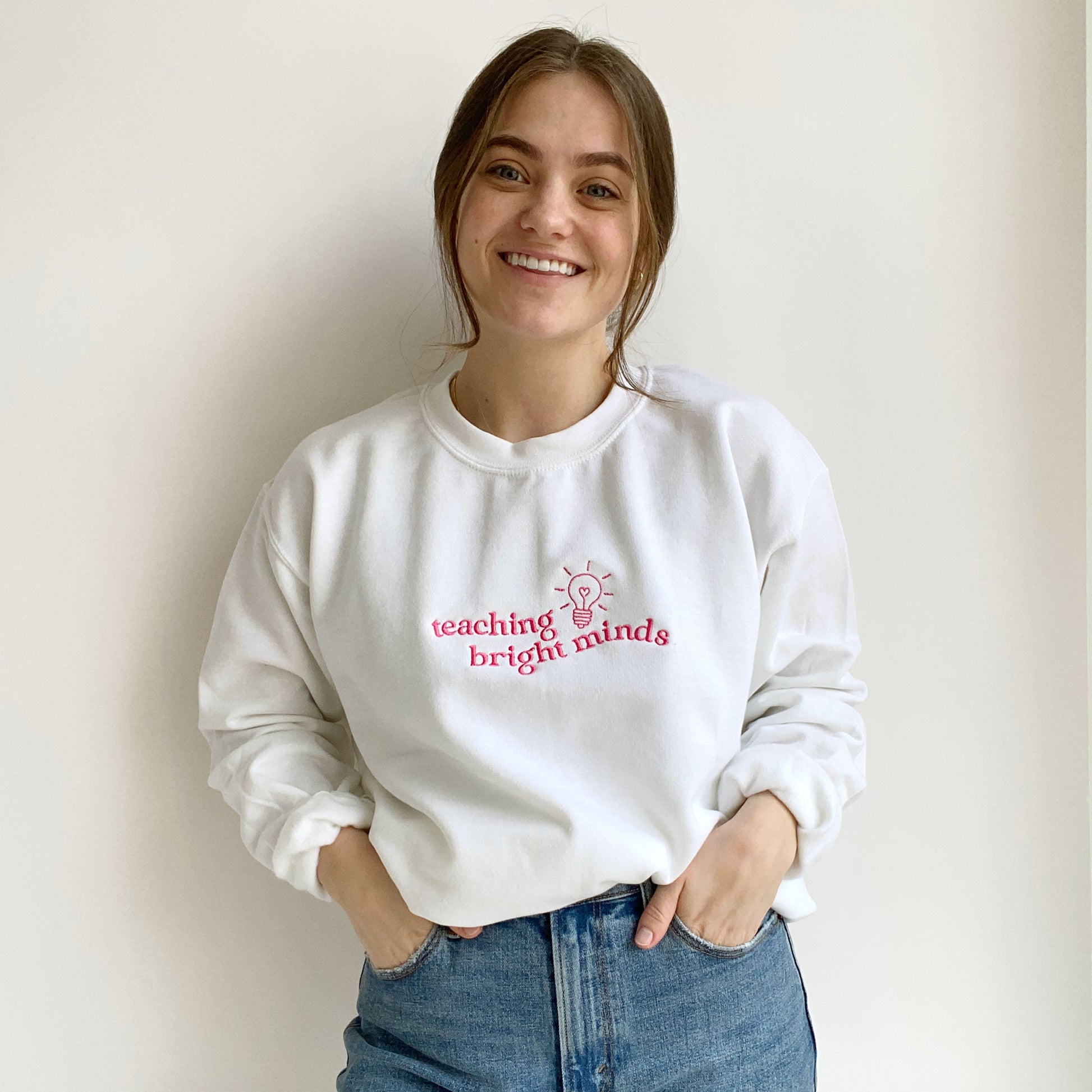 young woman wearing a white teacher pullover sweatshirt with a teaching bright minds lightbulb embroidered design on the chest