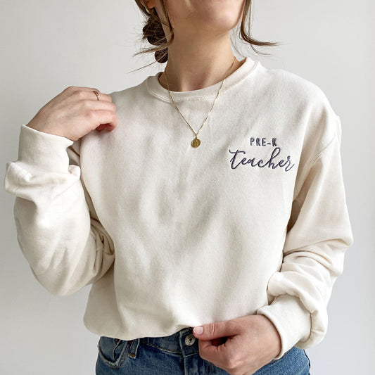 young woman wearing a sweet cream crewneck sweatshirt with custom teacher grade design embroidered in smoky orchid thread