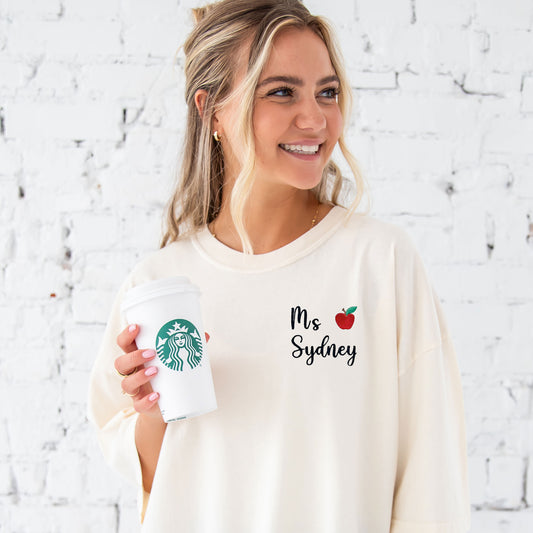 young woman wearing an ivory comfort colors t-shirt with embroidered name and mini apple on the left chest
