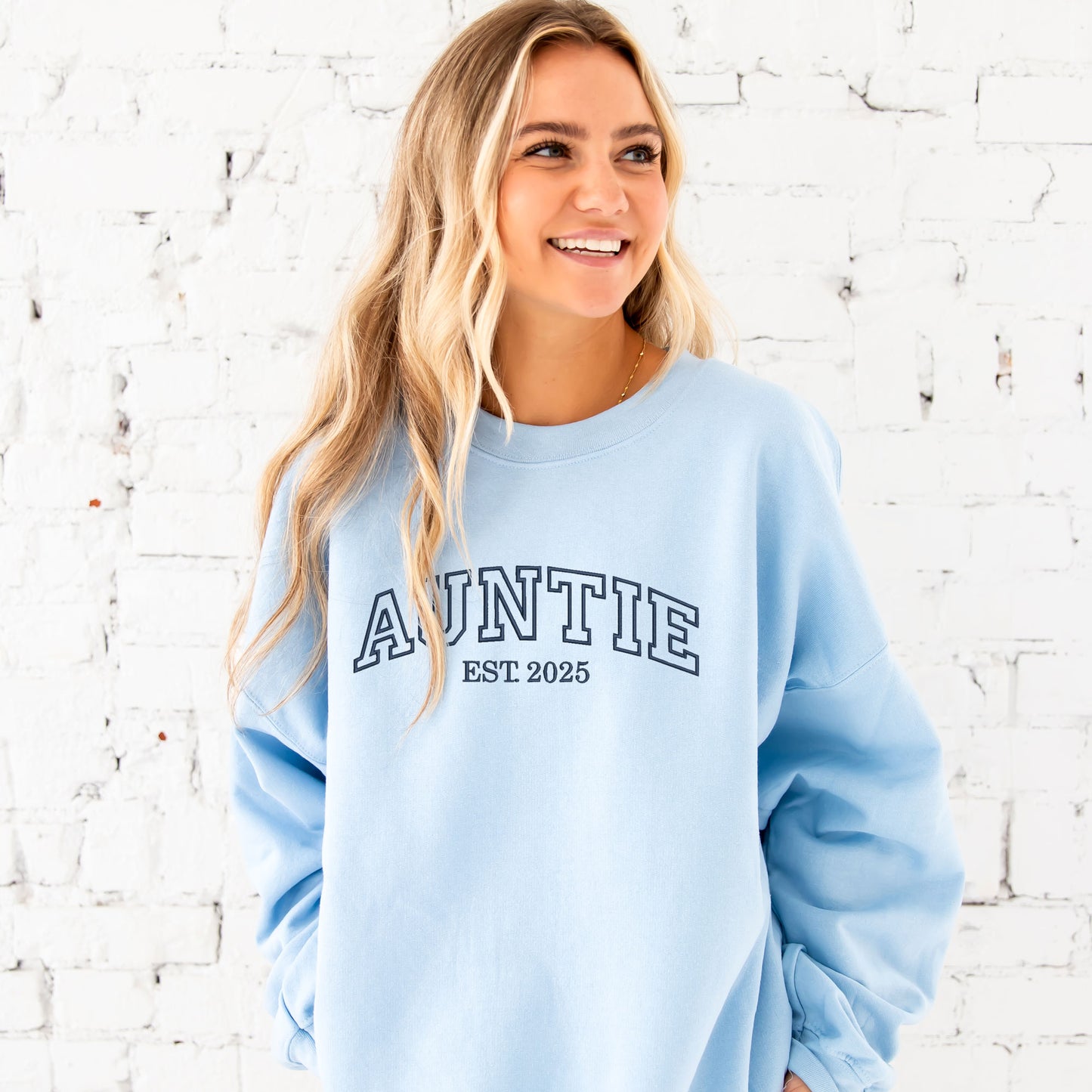 young woman wearing a light blue crewneck sweatshirt with embroidered AUNTIE in all caps athletic block font across the chest  and Est. 2025 underneath in navy thread.