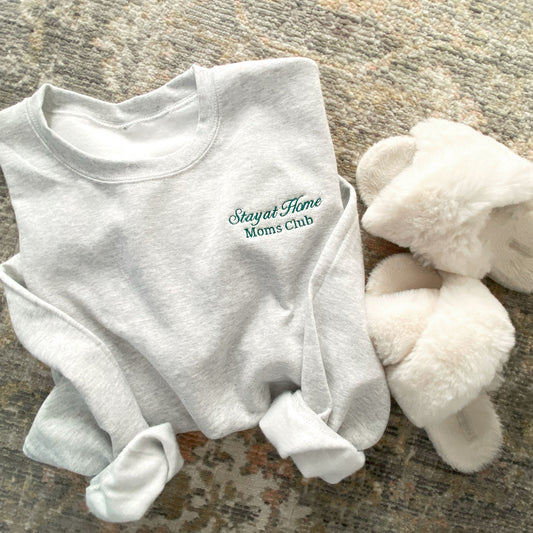 flat lay of an ash crewneck sweatshirt with embroidered stay at home moms cub design on the left chest in hunter green thread styled with fuzzy slippers