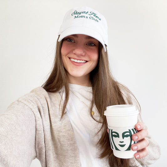 woman wearing a white hat with stay at home moms club embroidered in a dark green thread