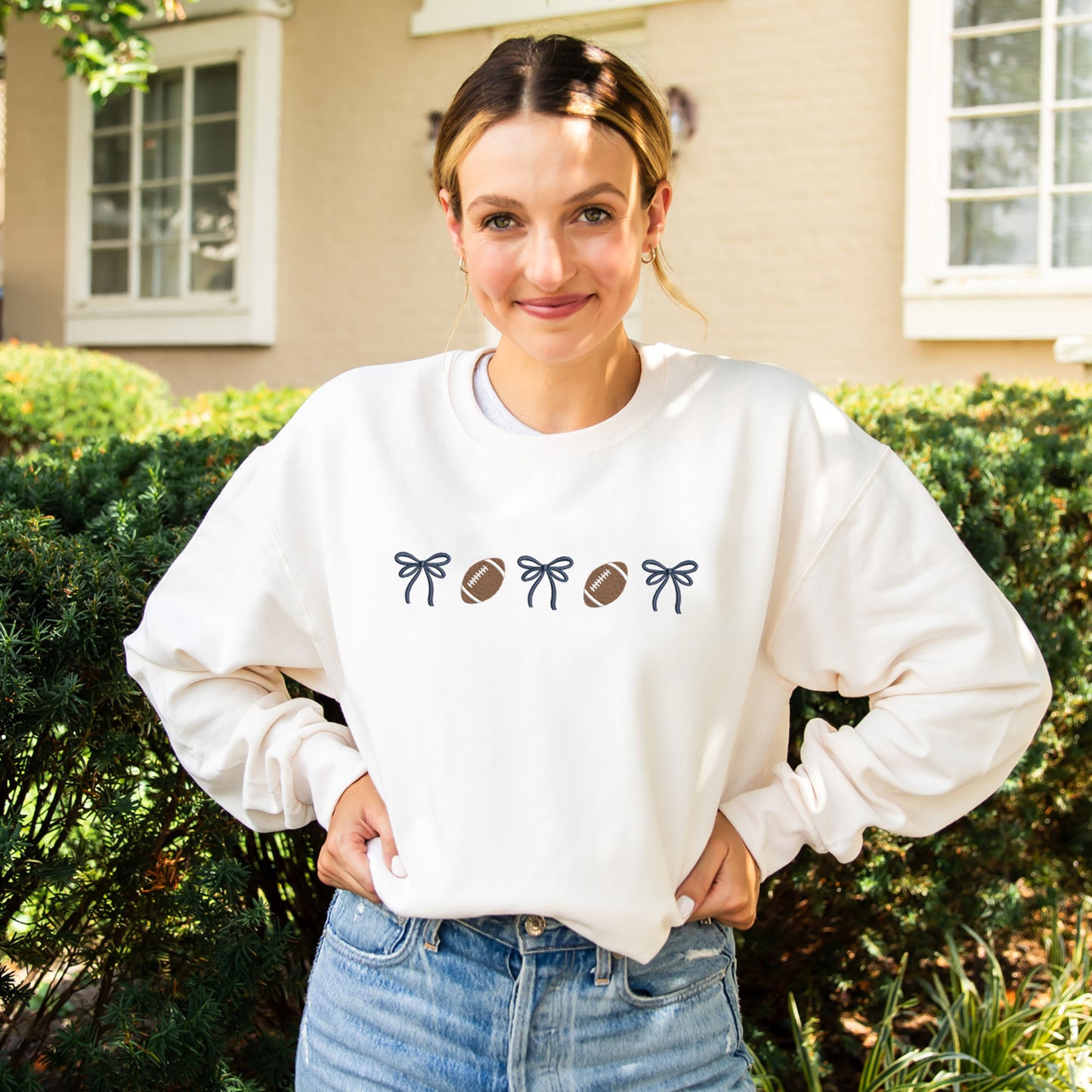 woman wearing a sweet cream crewneck sweatshirt with embroidered alternating bows and footballs design across the chest. The bow are embroidered in navy thread.