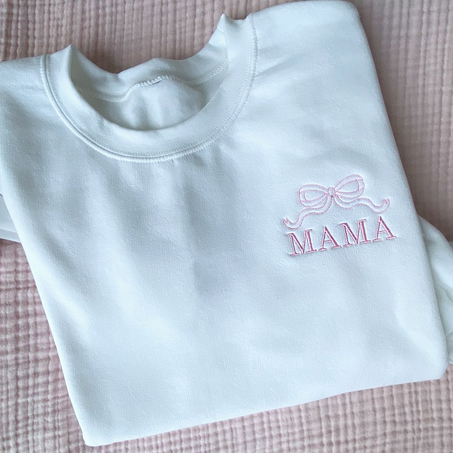 white crewneck sweatshirt with embroidered outline bow and all caps mama design on the left chest in baby pink thread