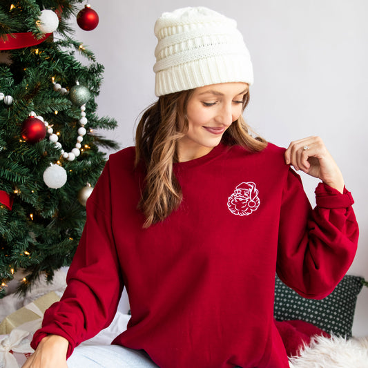 woman sitting by her christmas tree wearing an ivory knit beanie and a red crewneck sweatshirt with santa embroidered in a white thread on the left chest