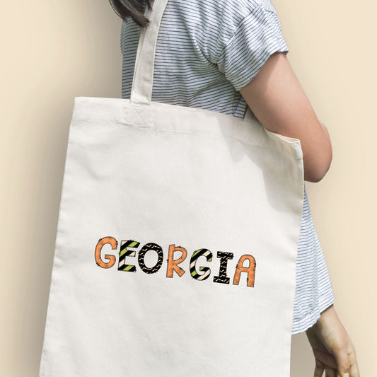 Girl holding a natural tote bag with a fun custom name print in a mixed halloween inspired pattern text.