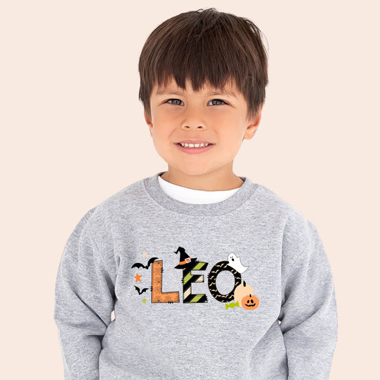 little boy wearing a heather gray crewneck sweatshirt with a custom name print in a mixed patterned Halloween text and accented with cute spooky icons like bats, a ghost, jack-o-lantern, and witch hat.