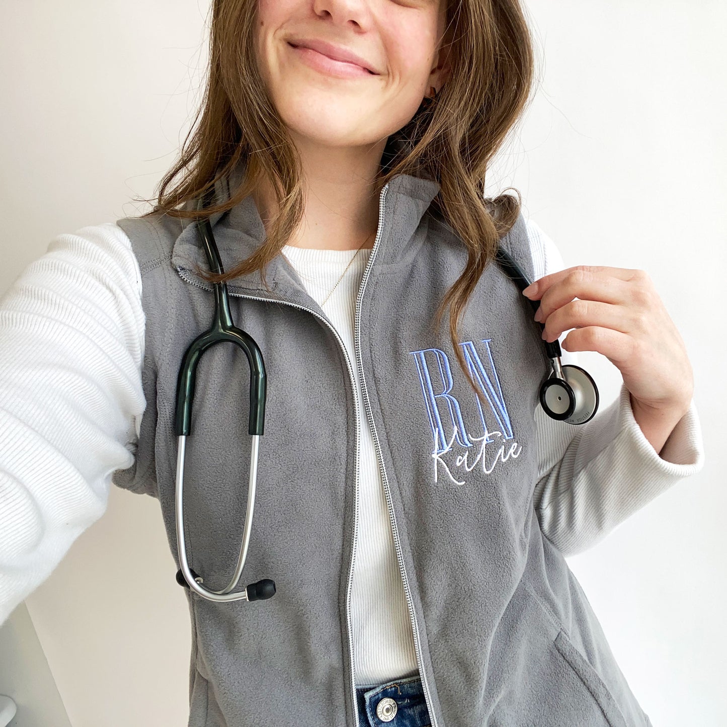 selfie of a young woman wearing a grey flecce vest with personalized RN embroidered design