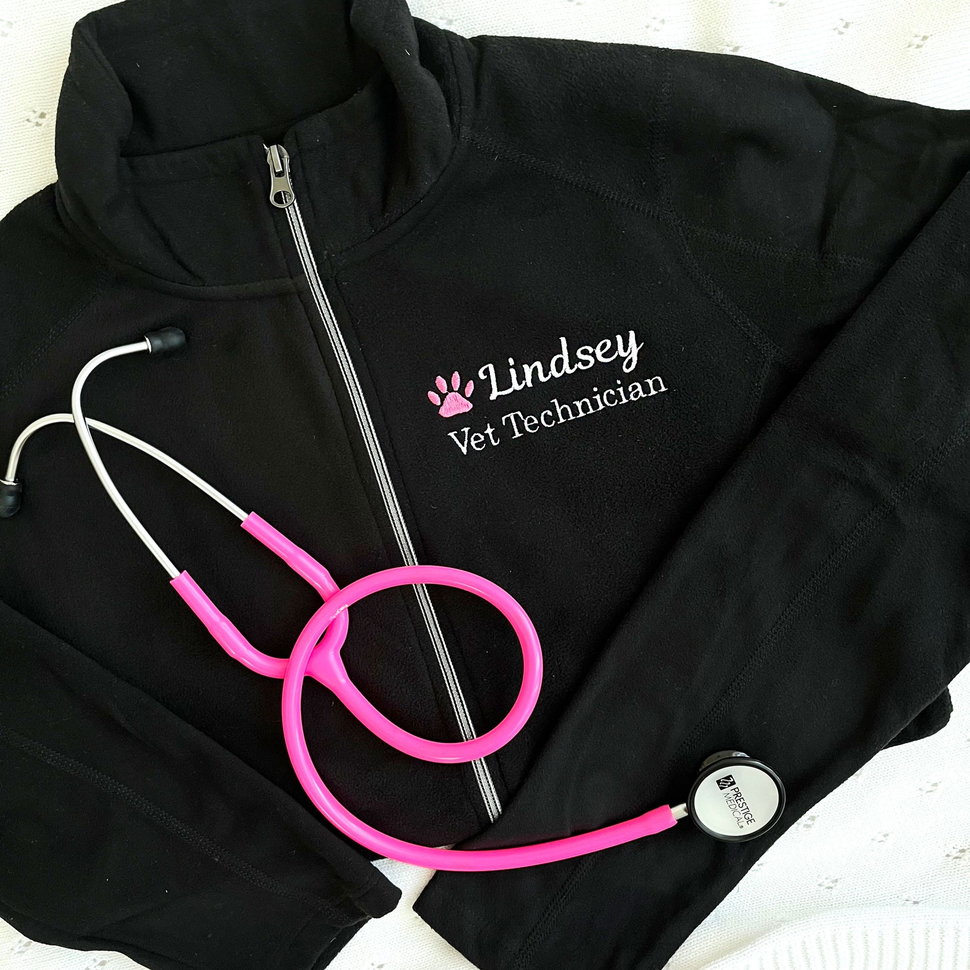 flat lay of a black full zip fleece jacket with embroidered pink paw print and name Lindsey with Vet Technician embroidered on the left chest in white thread