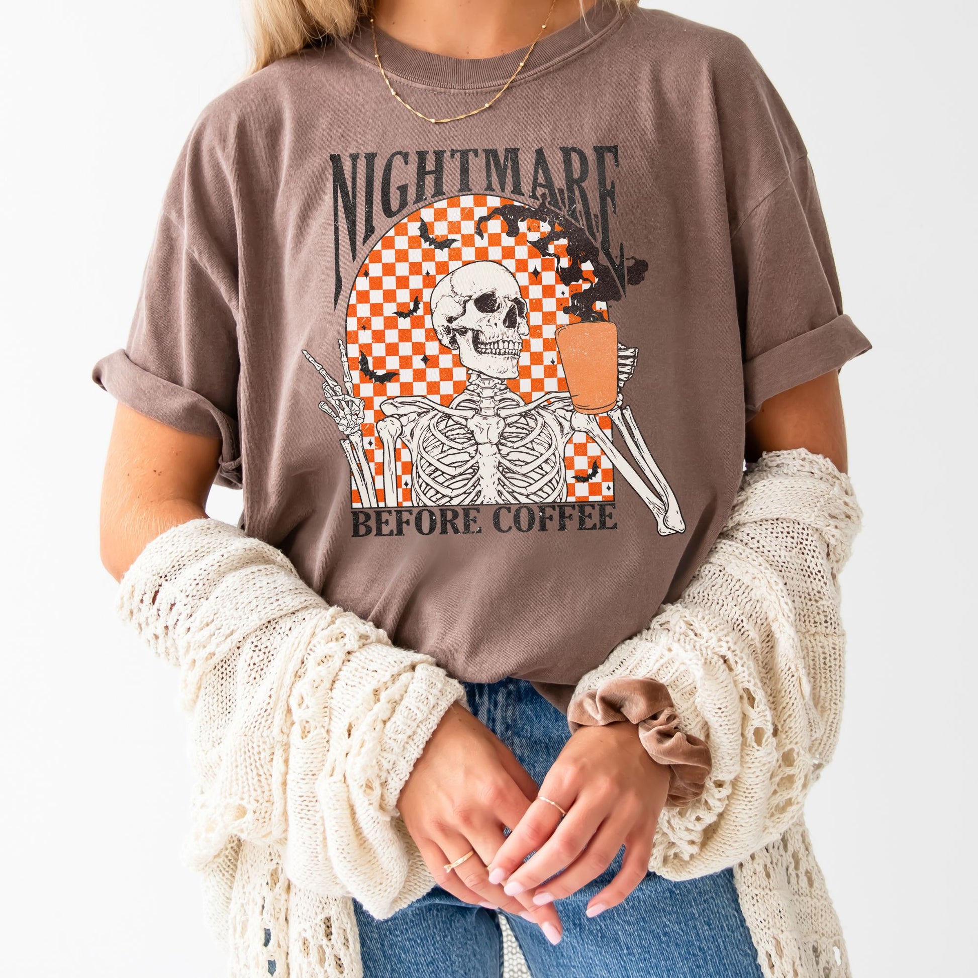young woman wearing an espresso  comfort colors t-shirt with a nightmare before coffee skeleton/bat print with a checkered background