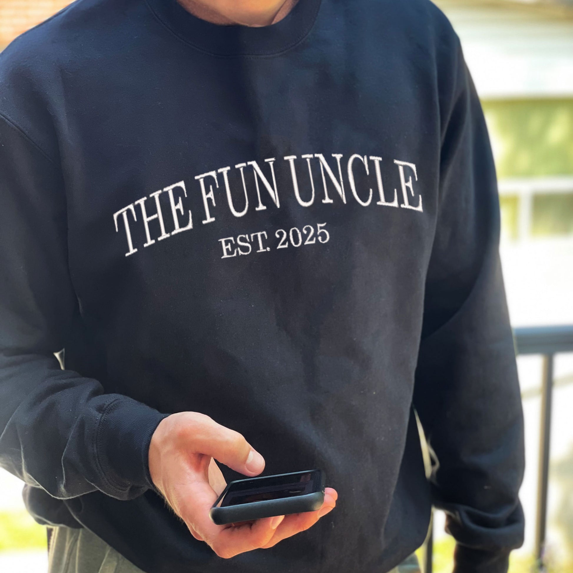 man wearing a black crewneck sweatshirt with embroidered The Fun Uncle est. 2025 in white thread large across the chest