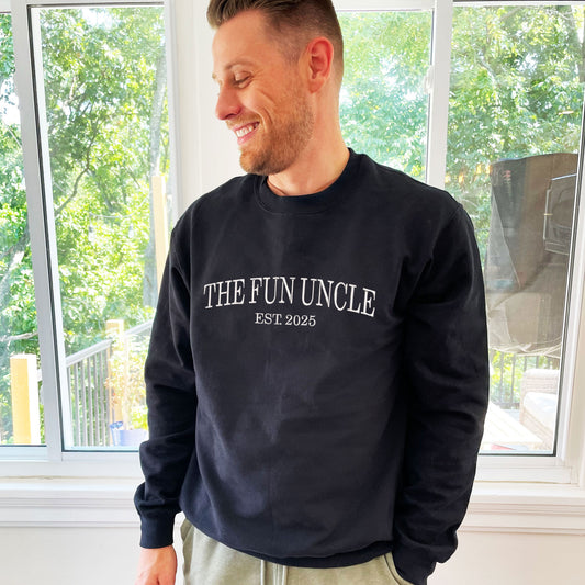 man wearing a black crewneck sweatshirt with embroidered The Fun Uncle est. 2025 in white thread large across the chest