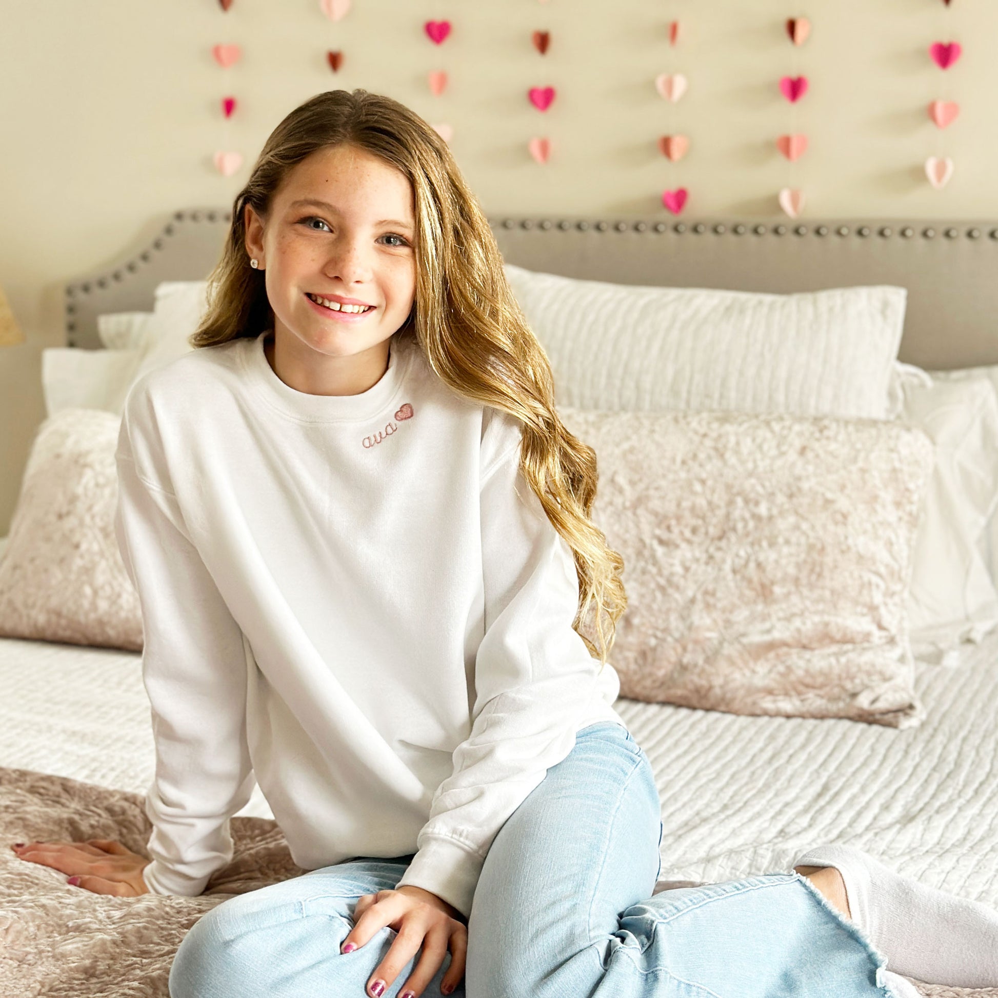 young girl modeling a white crewneck sweatshirt with her name embroidered along the neckline with a heart.